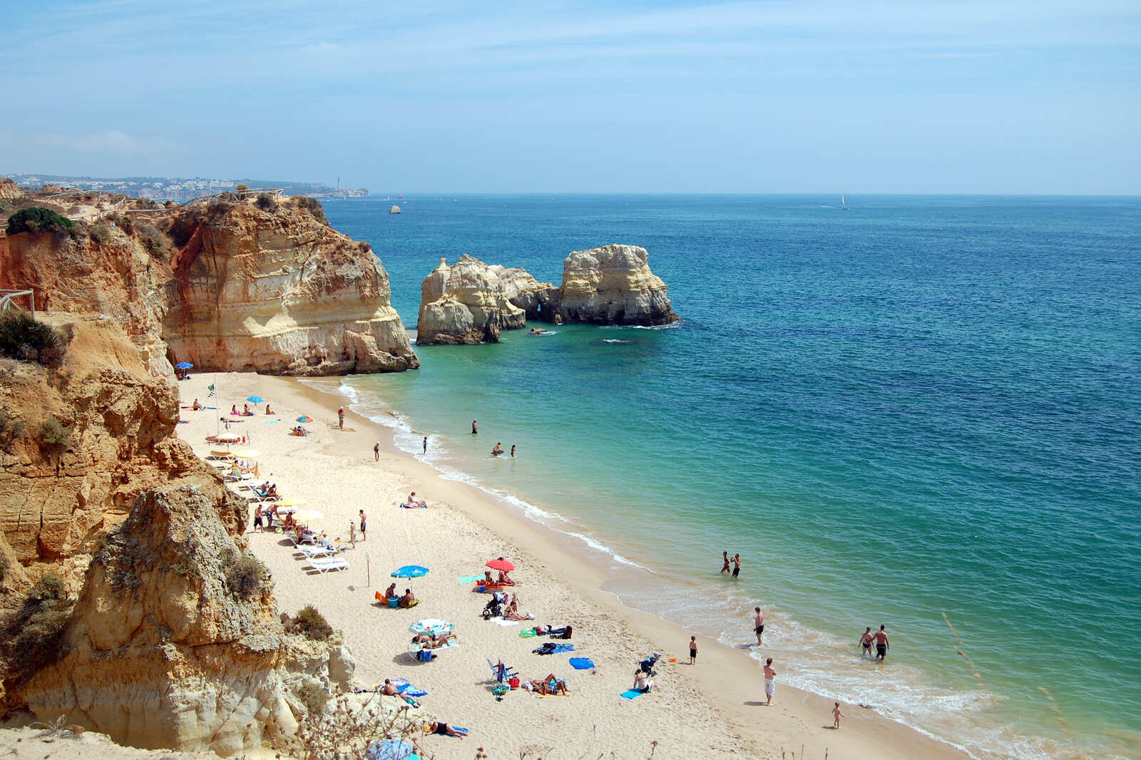 Fullsatt strand mellom oker klipper ved Praia da Rocha i Algarve i skarpt sommerlys